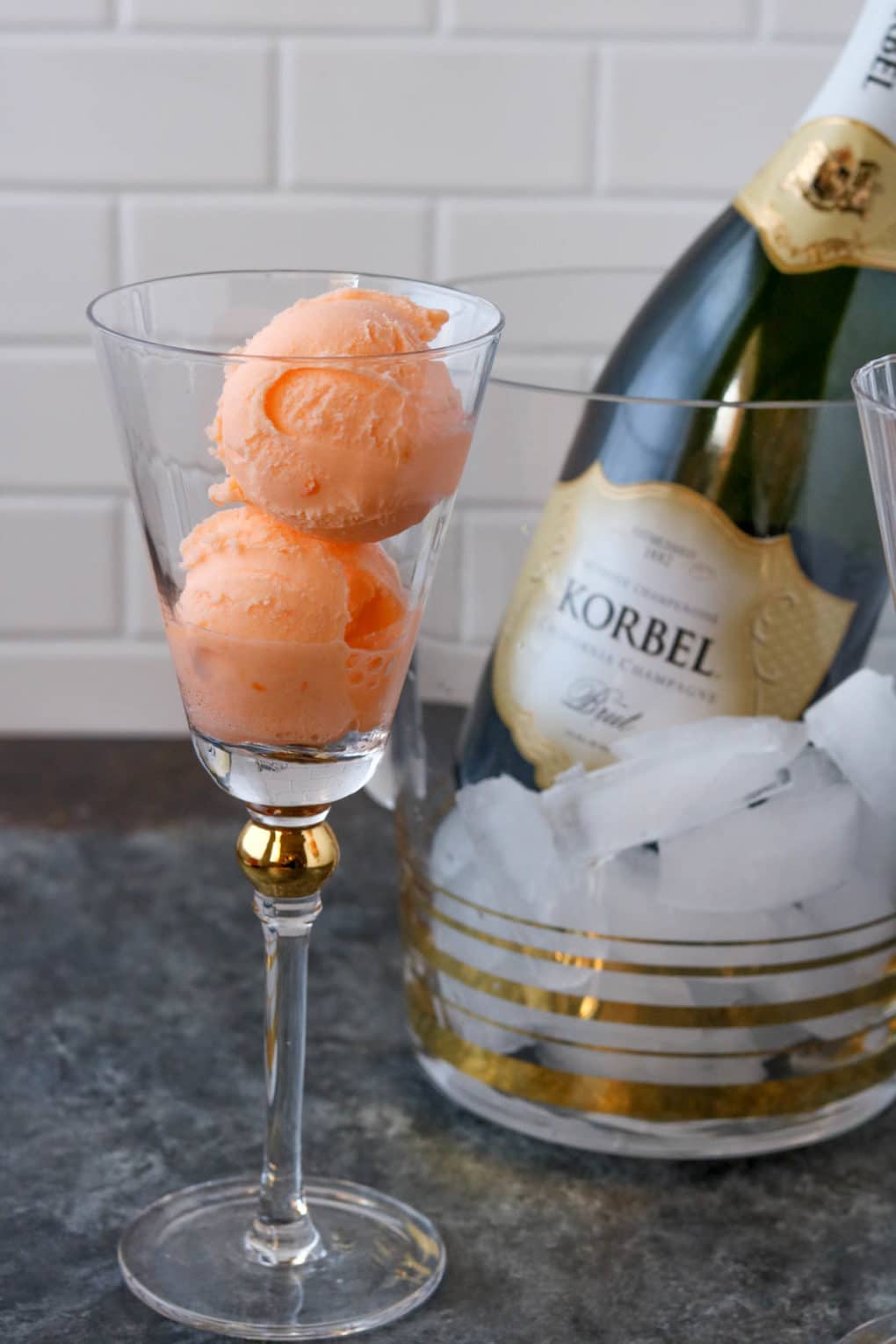 a champagne glass with two scoops of orange sherbet and a bottle of champagne in the background in a chill bucket