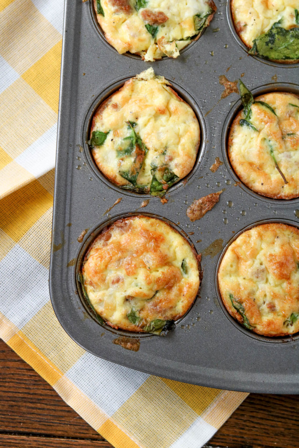A muffin tin with spinach sausage egg muffins. The silver muffin tin is sitting on a wood table with a yellow and white checkered towel underneath. 