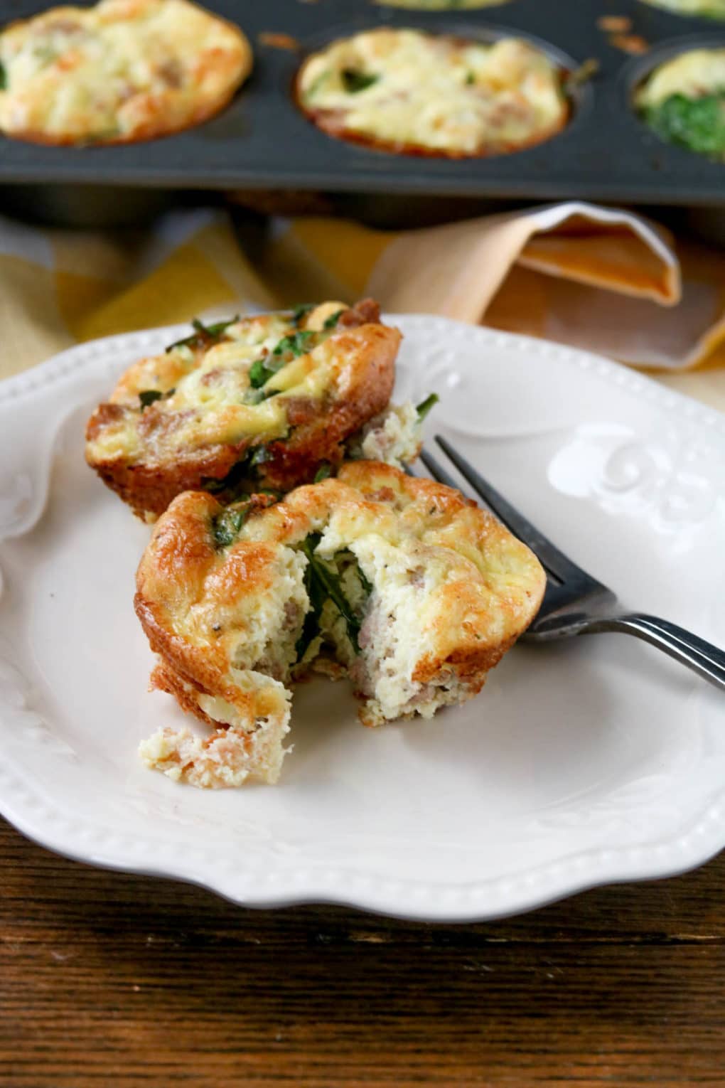 One spinach sausage egg muffin on a white plate with a silver fork. There is a muffin tin of more spinach sausage egg muffins in the background. 