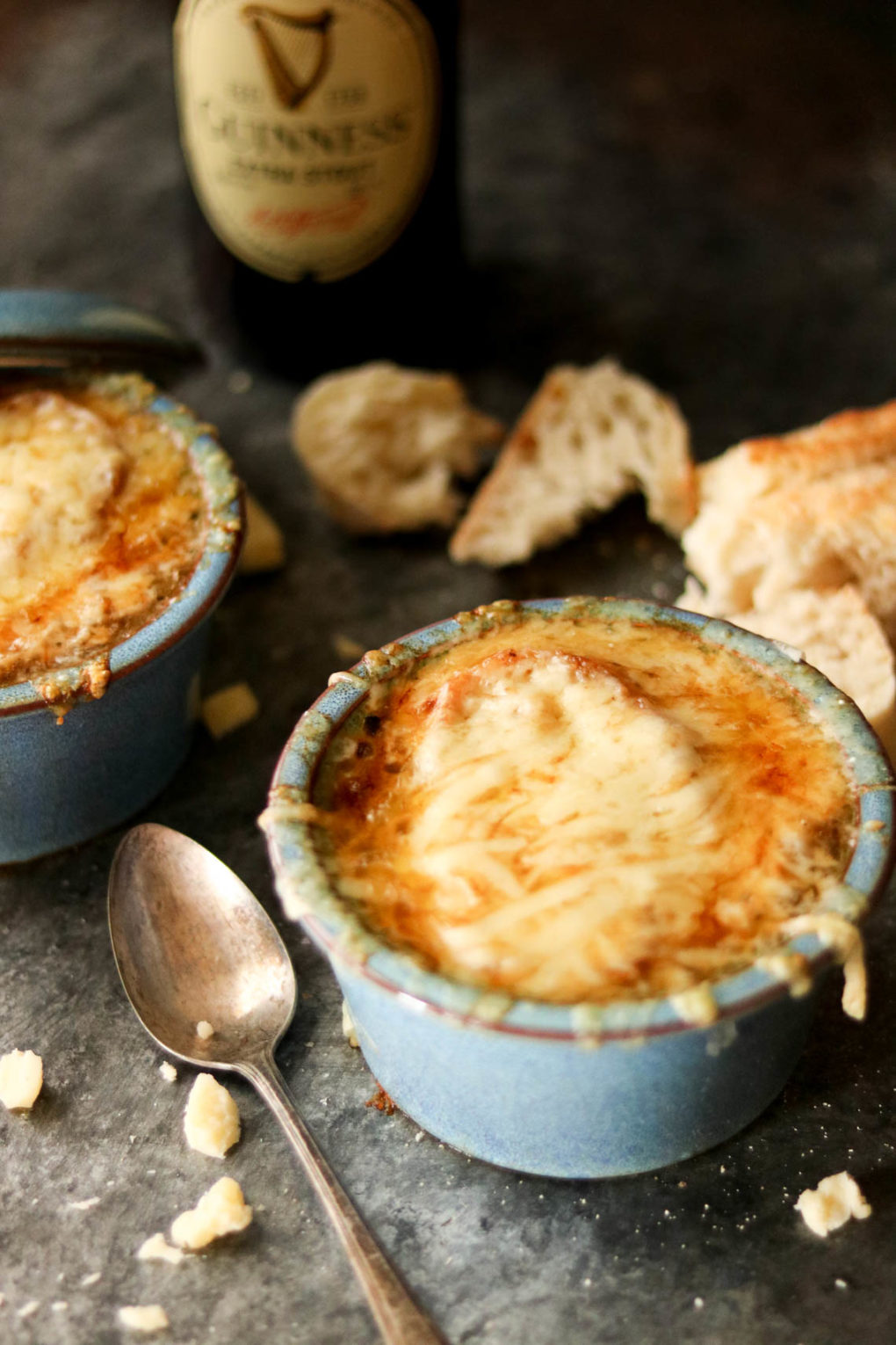 two bowls of Guinness french onion soup. there's a spoon laying nearby and a bottle of Guinness and a baguette in the background