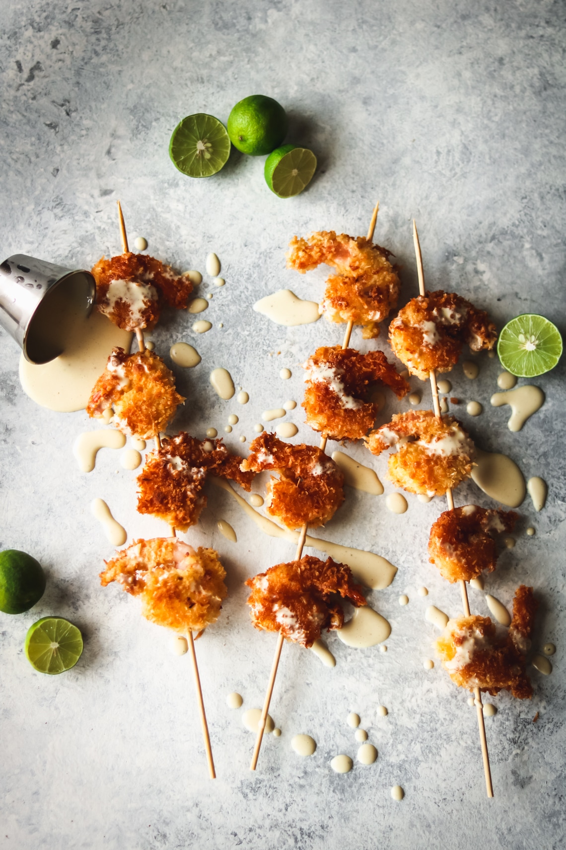 coconut shrimp on wooden skewers with key lime aioli drizzled all over. There are sliced key limes laying around the coconut shrimp on the table and a small sauce cup of the key lime aioli