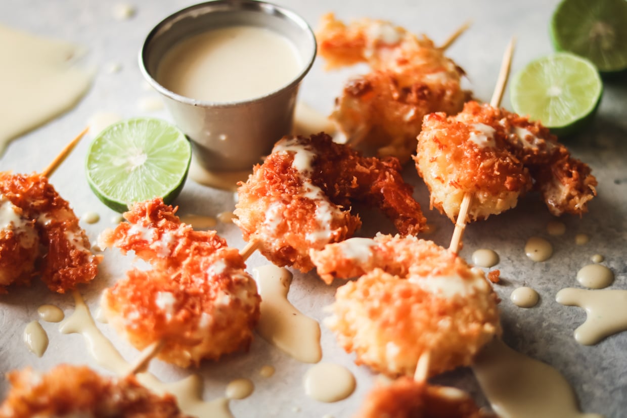 coconut shrimp on wooden skewers with key lime aioli drizzled all over. There are sliced key limes laying around the coconut shrimp on the table and a small sauce cup of the key lime aioli