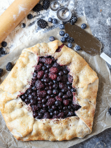 a rustic berry galette sitting on a table - there is flour, measuring spoons, and a rolling pin sitting nearby