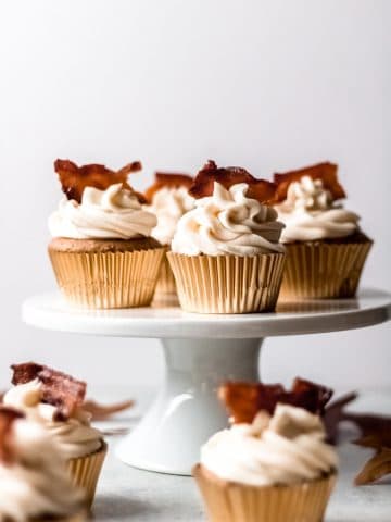4 maple bacon spice cupcakes on a cake stand with additional cupcakes on the table surrounding the cake stand