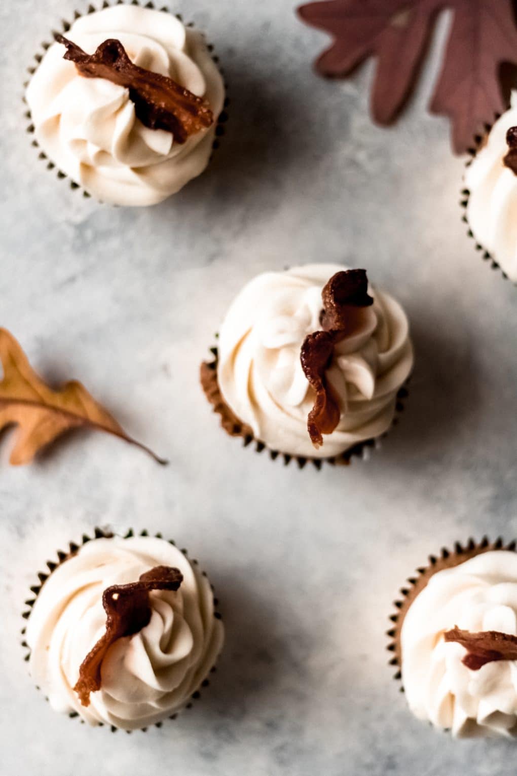 4 maple bacon cupcakes on a table with fall leaves laying nearby