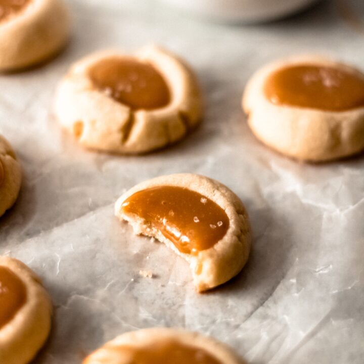 four salted caramel thumbprint cookies on a table with one having a bite out of it and a bowl of caramel sitting in the background