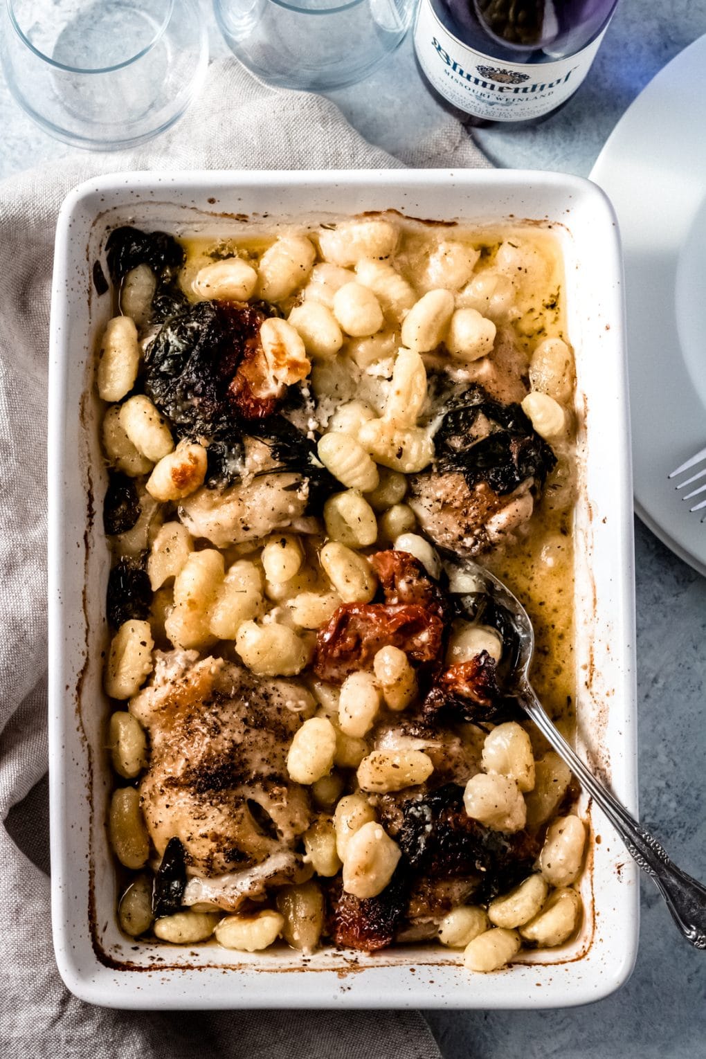 a 9x13 dish of baked chicken gnocchi with a silver serving spoon in the dish. There are plates, two wine glasses and a bottle of wine surrounding the dish.