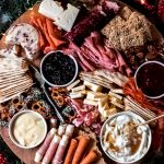 A round wooden holiday charcuterie board covered with various meats, cheese, crackers, and sweets, surrounded by holiday greenery