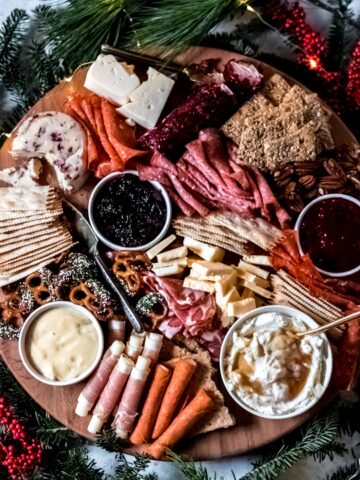 A round wooden holiday charcuterie board covered with various meats, cheese, crackers, and sweets, surrounded by holiday greenery