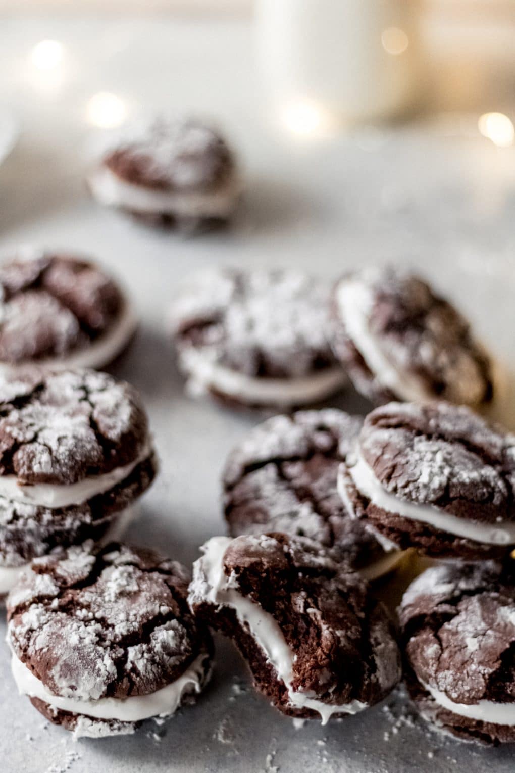 several hot cocoa sandwich cookies on a table with white christmas lights and a jug of milk in the background