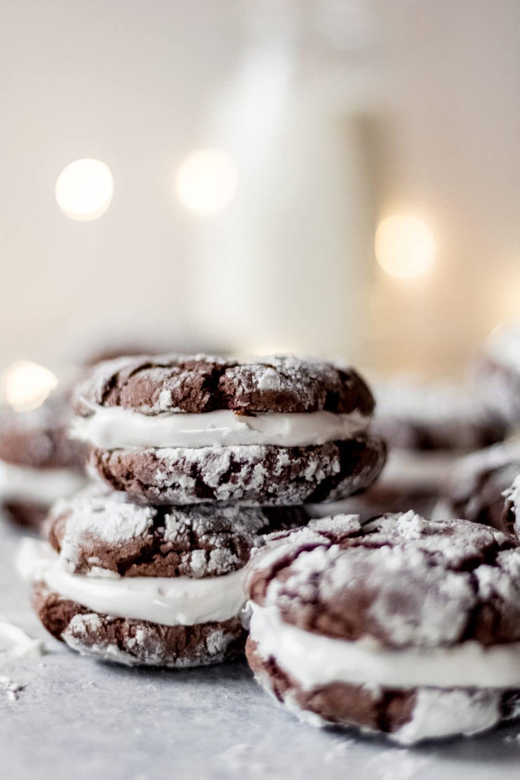 Three hot cocoa sandwich cookies stacked with additional cookies in the background. There's white christmas lights and a jug of milk in the background as well.