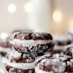 Three hot cocoa sandwich cookies stacked with additional cookies in the background. There's white christmas lights and a jug of milk in the background as well.