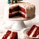 A red velvet cake with two layers on a white cake stand. Two slices have been cut out and served onto plates. Two gold forks sit next to the plates. There are tiny green and gold bottle brush trees on top of the cake and white lights glisten in the background.