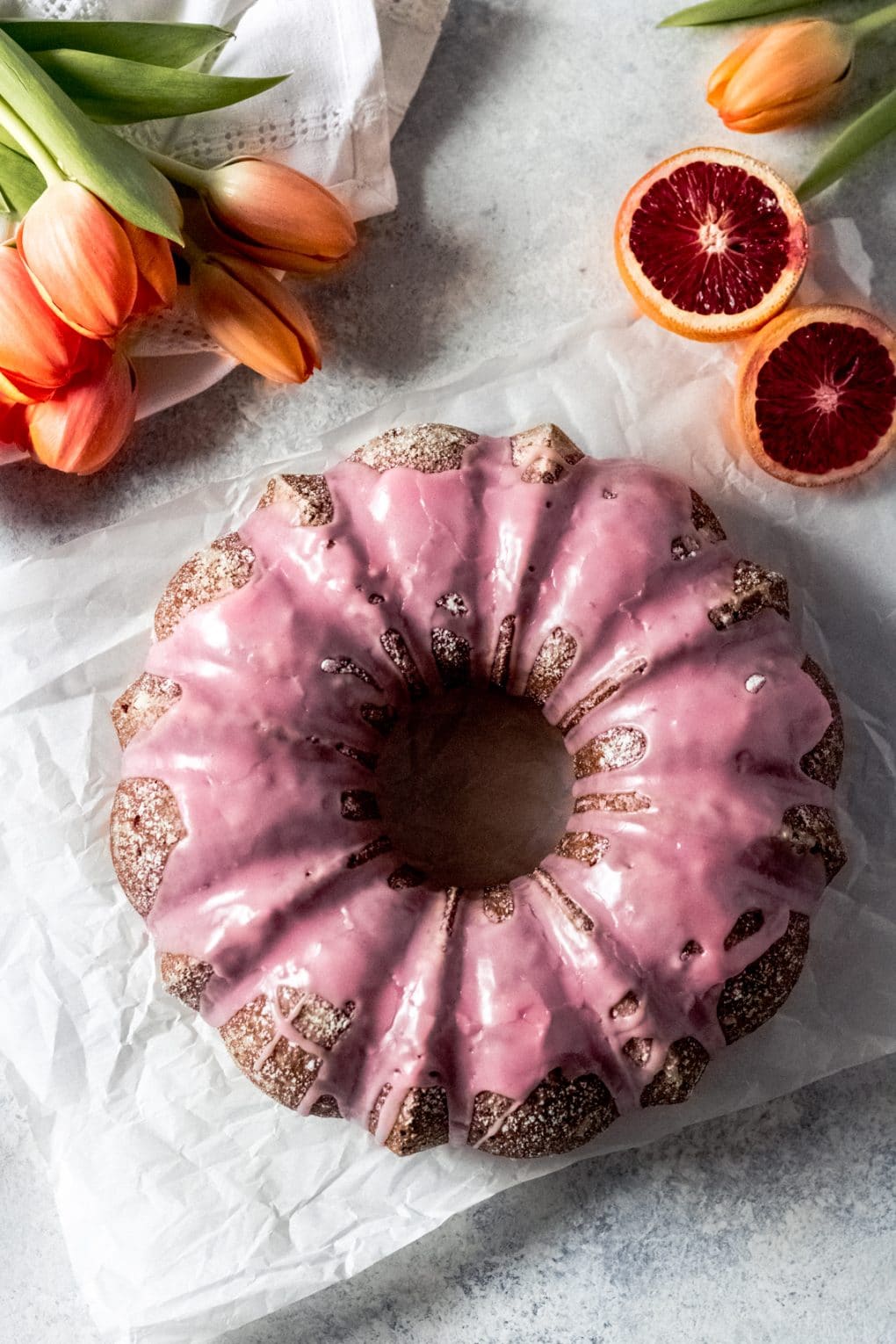 an overhead shot of a blood orange olive oil cake with a pink blood orange glaze on top. There are blood oranges in the upper right corner and orange tulips in the upper left.