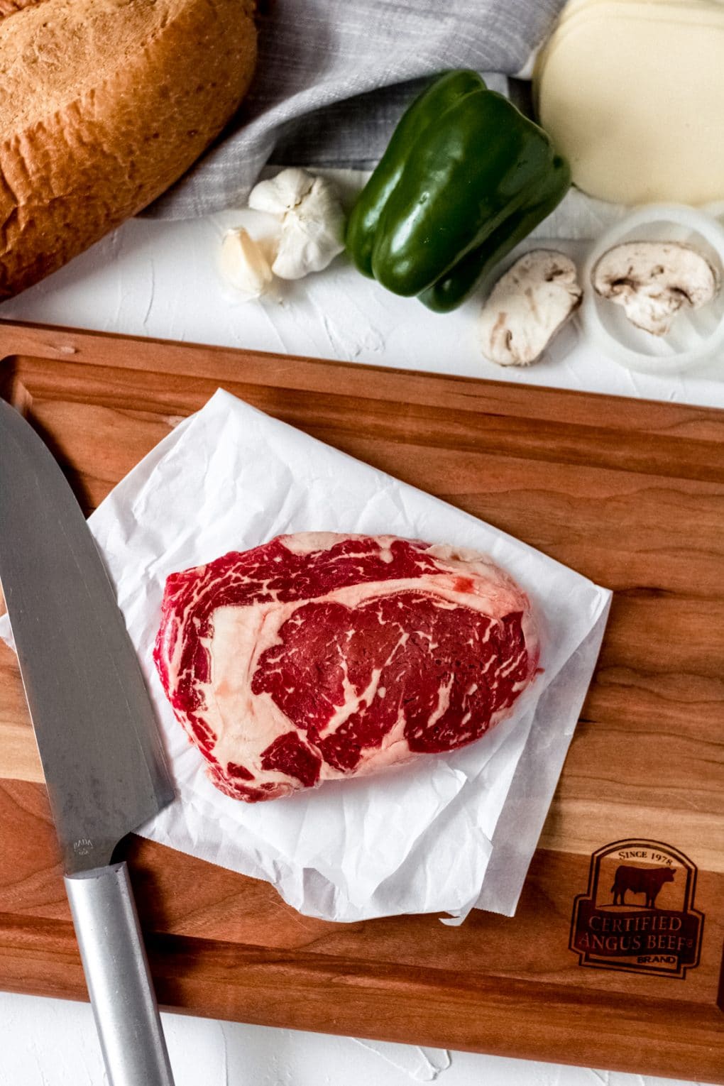 a certified angus beef brand ribeye steak on a cutting board. There's a chef knife to the left and the ingredients for the philly cheesesteak stuffed french bread are laying on the table above the cutting board