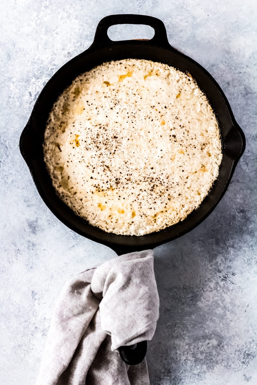 baked ricotta dip in a cast iron skillet with a neutral colored towel wrapped around the handle.