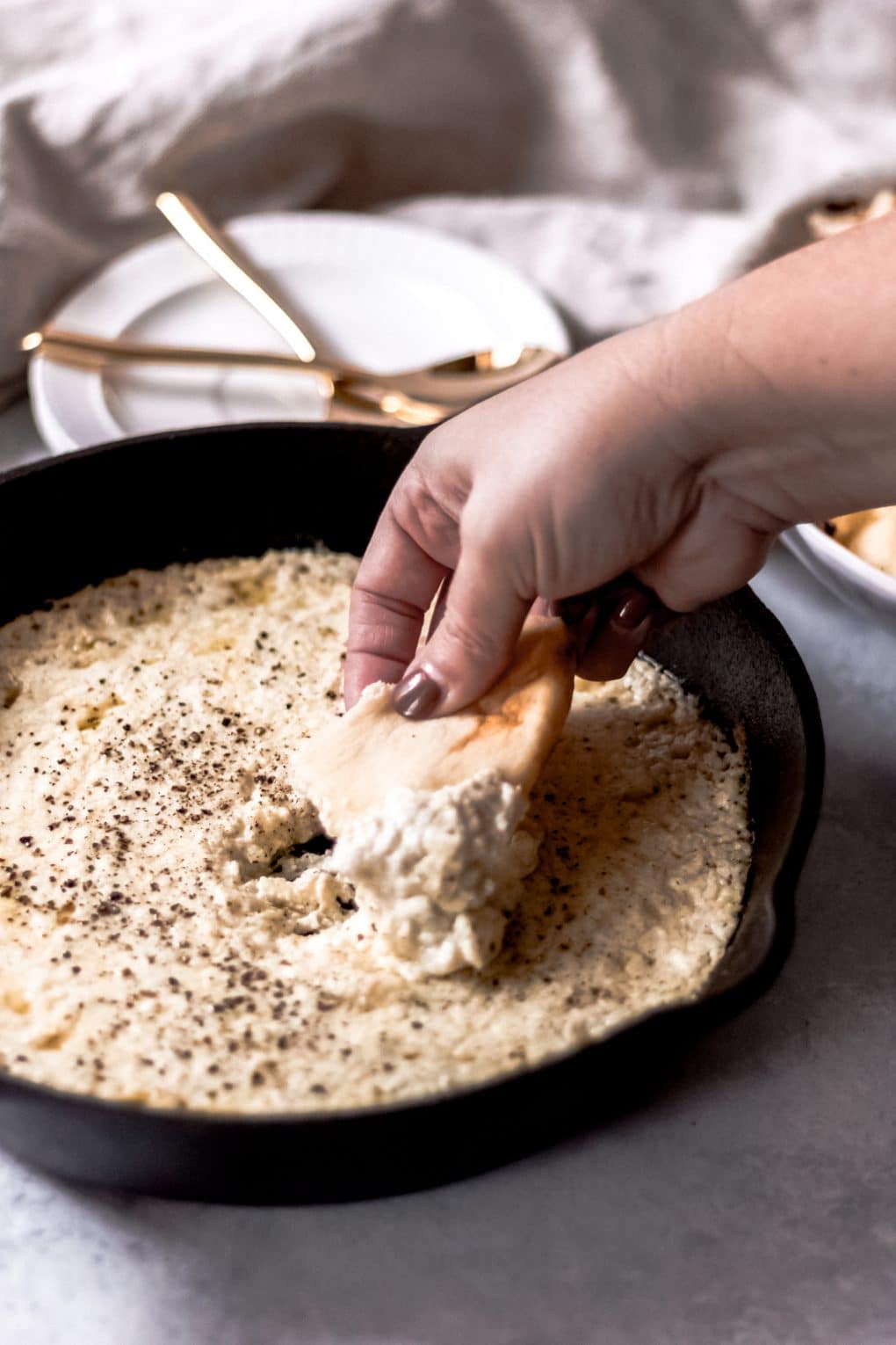 A woman dipping a piece of pita bread into a cast iron skillet of baked ricotta dip There are two white plates and two gold forks in the background and a neutral-colored towel.