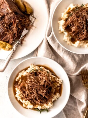 Two white bowls with mashed potatoes topped with shredded Mississippi pot roast. The Mississippi pot roast is on a white serving platter with a silver carving knife to the left of the bowls.