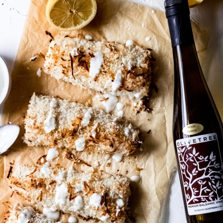 four coconut crusted Mahi-Mahi-Mahi fillets on a brown sheet of parchment paper. They have a creamy lemon sauce drizzled over the top of them and there's a bottle of Olive Tree's lemon balsamic vinaigrette laying next to them on the table.