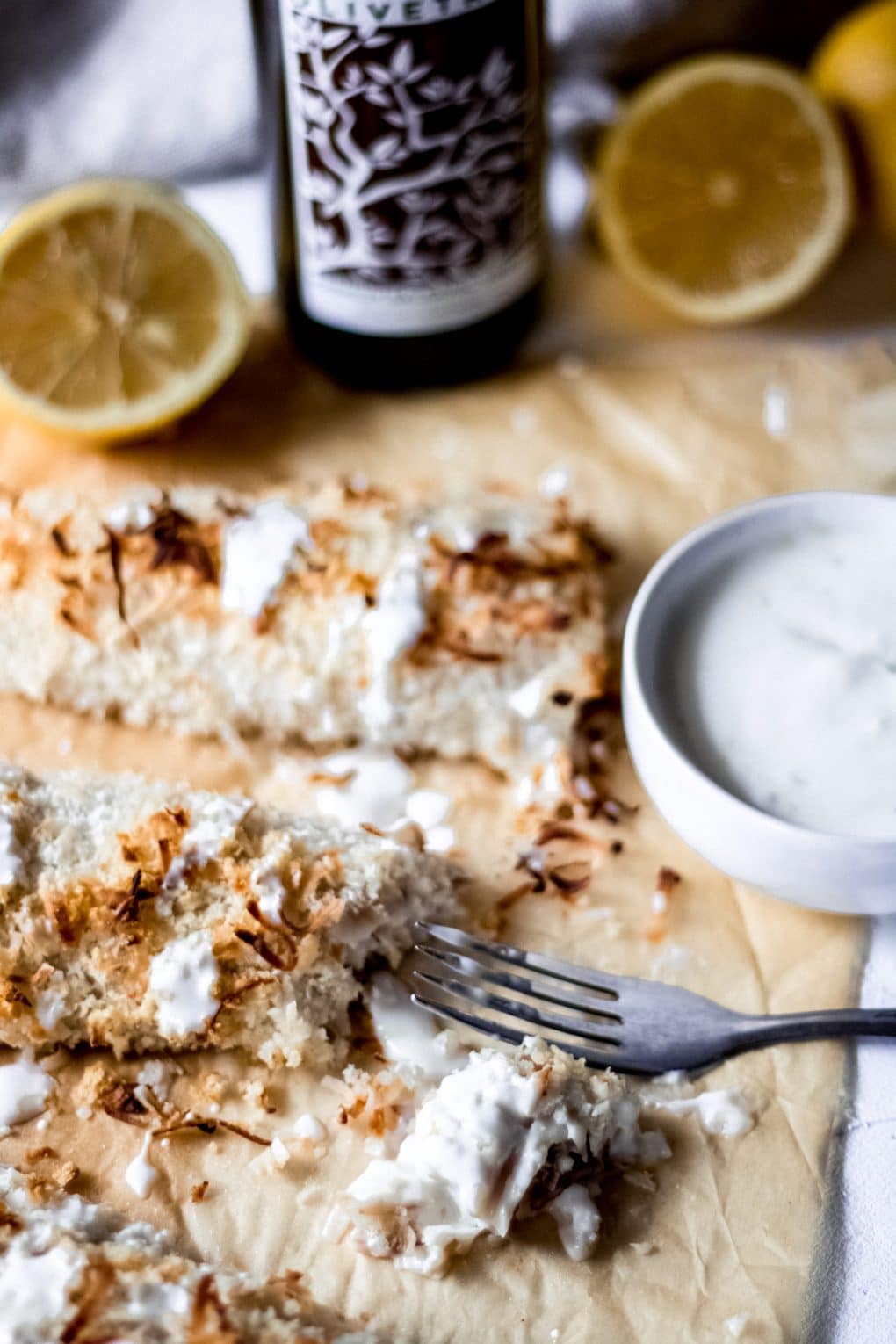 two fillets of coconut crusted Mahi-Mahi on brown parchment paper with creamy lemon sauce drizzled. There's a fork and a bit has been cut out of one fillet. 