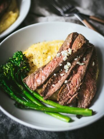 A white bowl with slices of skirt steak sitting on a bed of blue cheese polenta with broccolini on the side