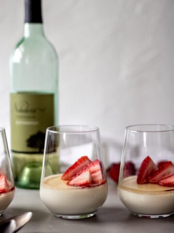 three glasses of vanilla panna cotta with white wine sauce with fresh strawberries in each glass. There's a bottle of white wine in the background and spoons laying in front of the glasses.