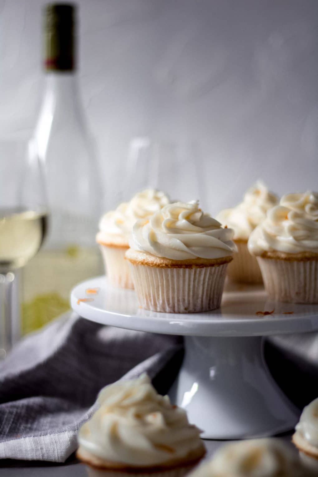 orange moscato cupcakes sitting on a white cake stand, there are also a few cupcakes sitting on the table. In the background there is a bottle of moscato and two wine glasses. 