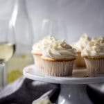 orange moscato cupcakes sitting on a white cake stand, there are also a few cupcakes sitting on the table. In the background there is a bottle of moscato and two wine glasses.