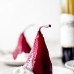 two red wine pears sit on white plates with a small dollop of coconut cream. There is a bottle of red wine in the background.