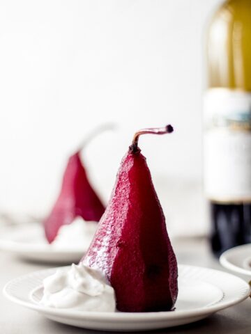 two red wine pears sit on white plates with a small dollop of coconut cream. There is a bottle of red wine in the background.