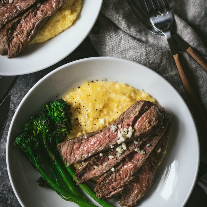 A white bowl with slices of skirt steak sitting on a bed of blue cheese polenta with broccolini on the side