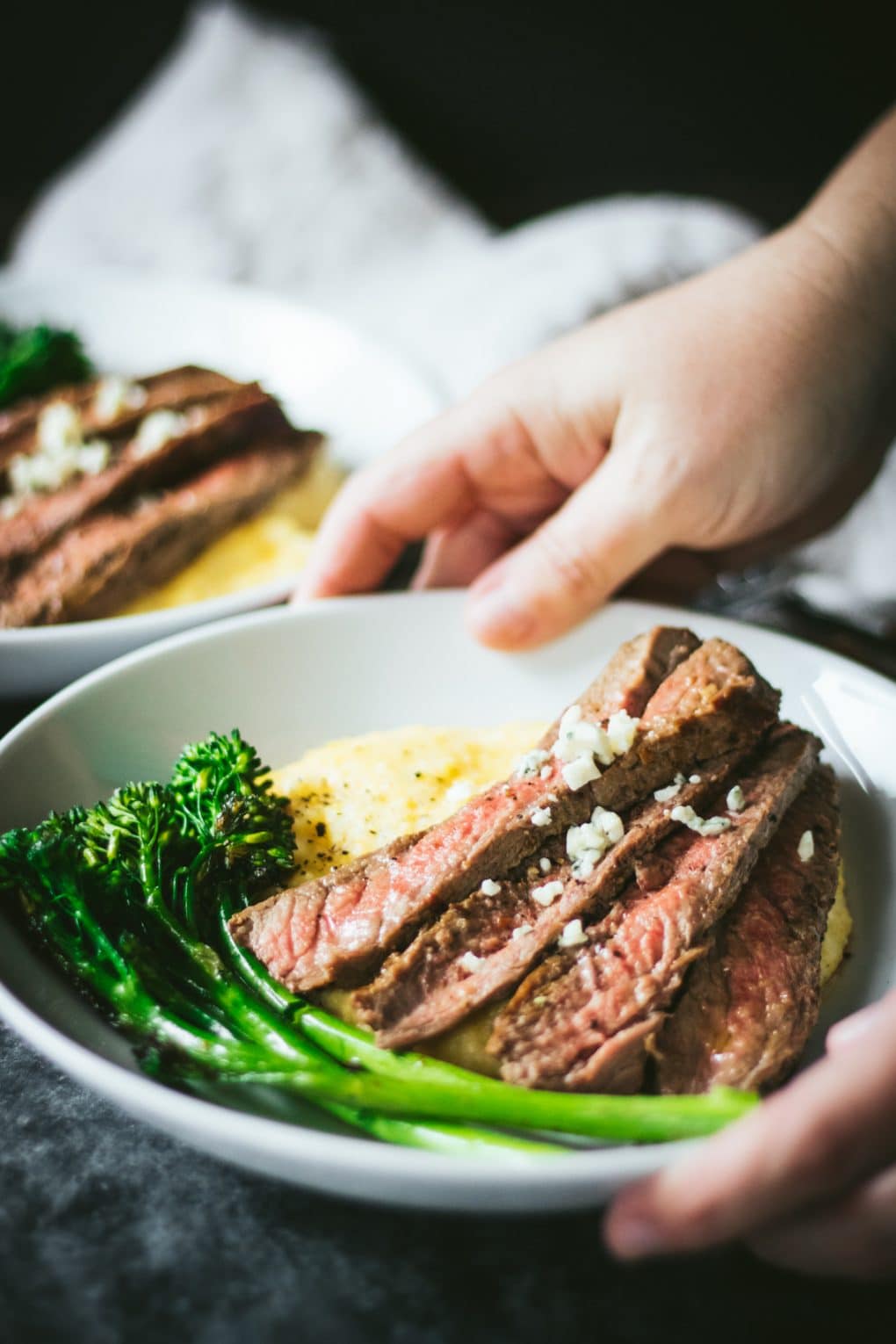 a woman serving a white bowl of skirt steak with blue cheese polenta and broccolini