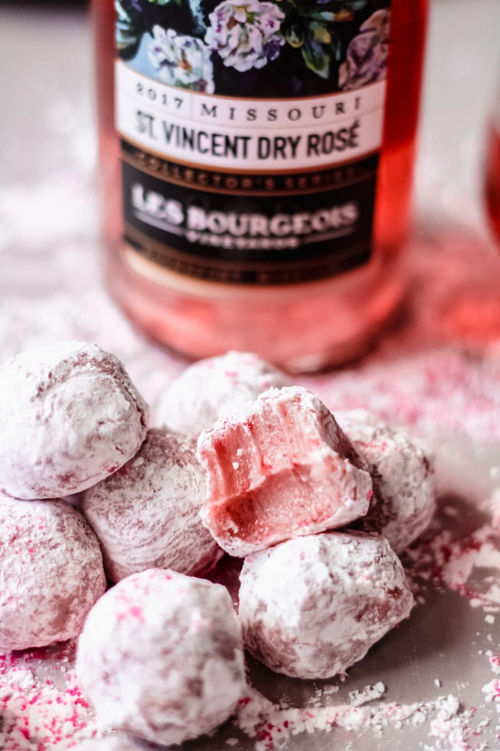 a pile of white chocolate rosé truffles on a table, there is one with a bite taken out of it and a bottle of rosé in the background
