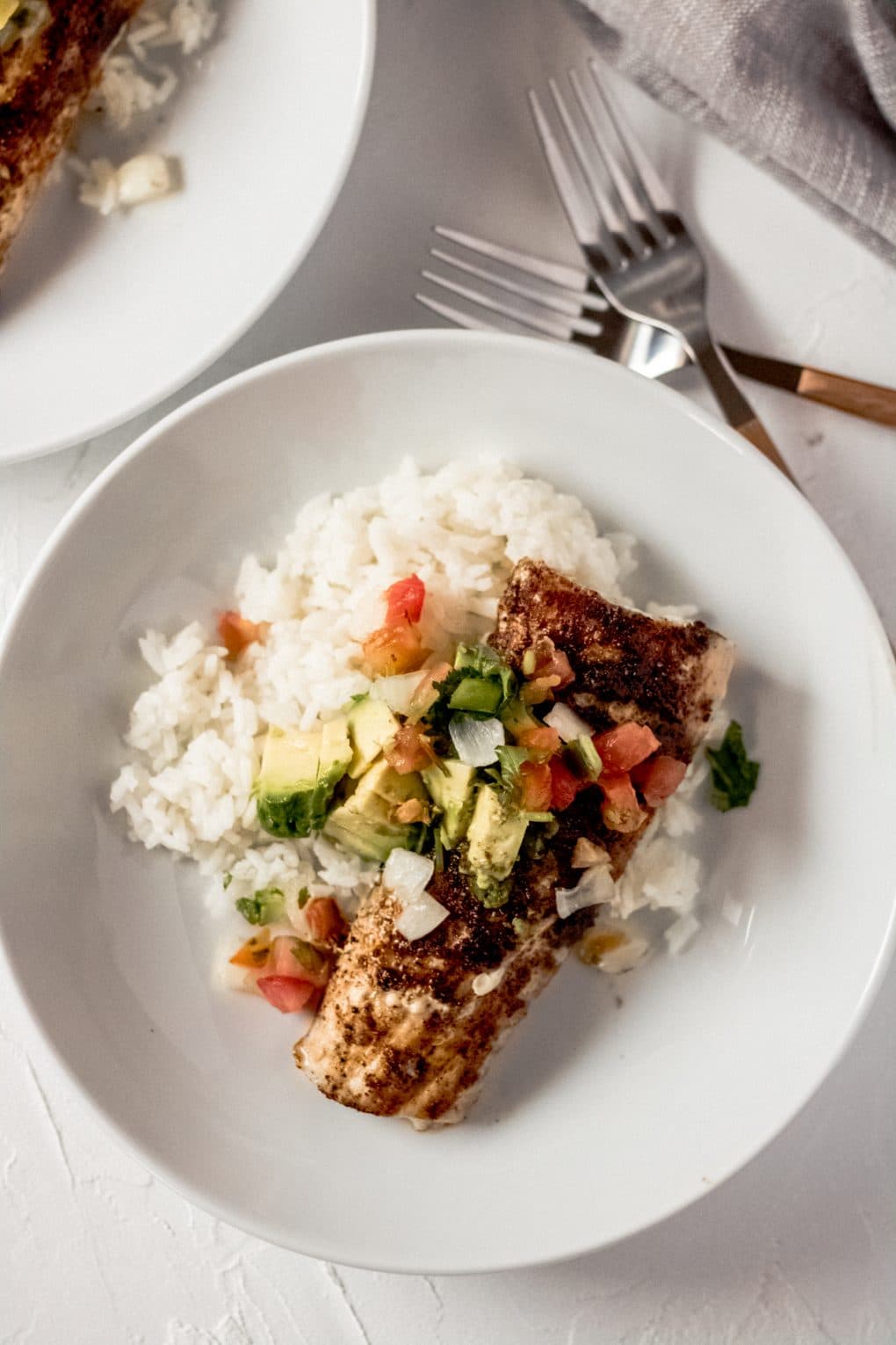 a grilled mahi mahi fillet on a bed of white rice in a white bowl topped with sliced avocado and pico de gallo.