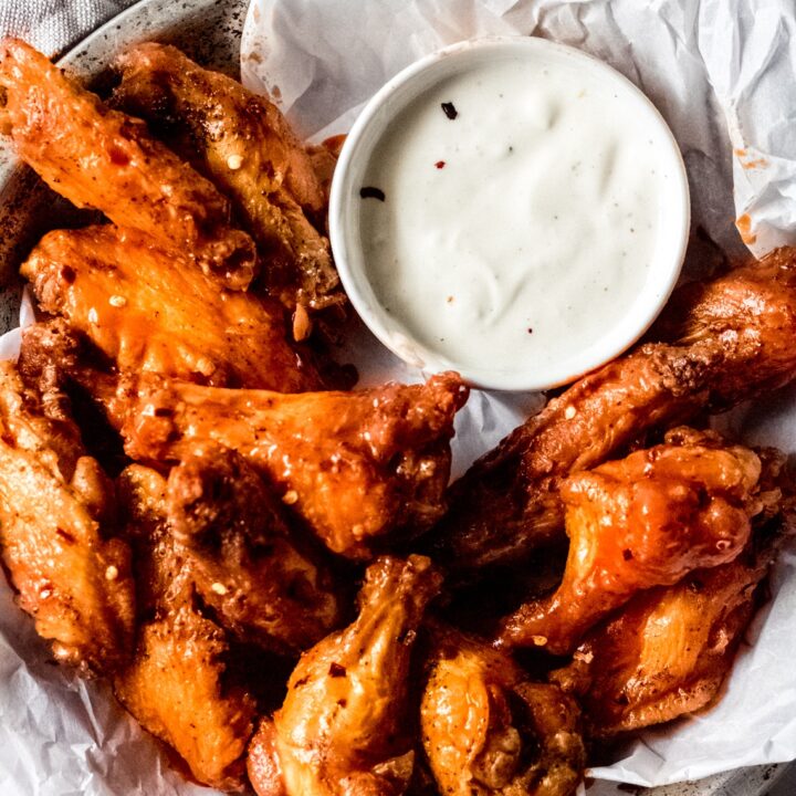 a bowl of hot spicy chicken wings sitting on white parchment paper and a bowl of blue cheese sauce