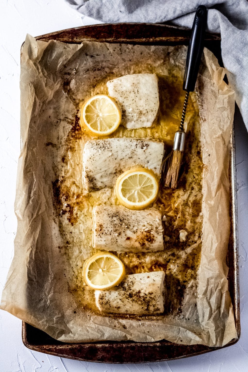 a sheet pan covered in parchment paper with four Mahi Mahi fillets, melted butter, slices of lemon, and a basting brush