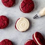 red velvet whoopie pies on a table, one has just been piped with icing and the piping bag is laying nearby