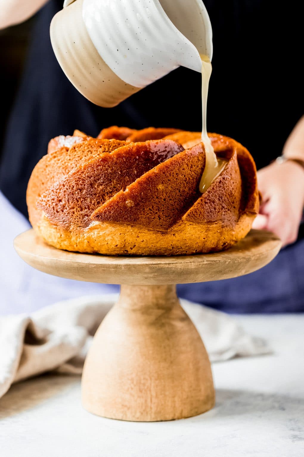 a woman pouring a sugar rum glaze over a baked rum cake on a cake stand